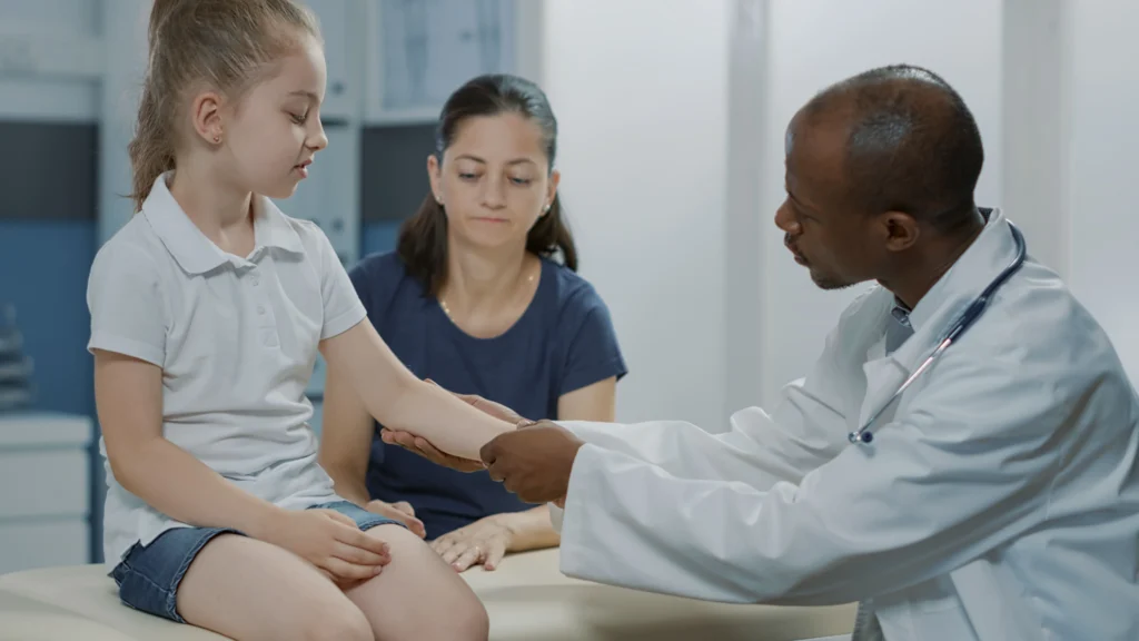 A doctor in a white coat examining a young girl's injured arm while her mother looks on in a medical office, representing medical treatment for a California child injury claim handled by Protection Law Group.