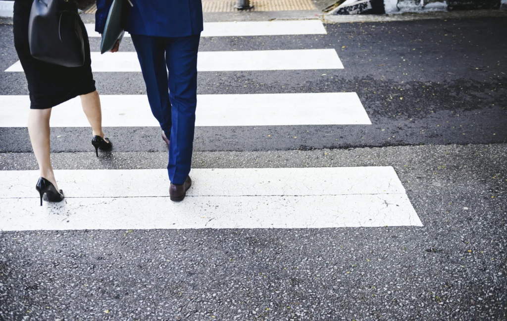 Lower-body view of two pedestrians in business attire walking across a painted white crosswalk on a city street, representing a California pedestrian accident claim handled by Protection Law Group.